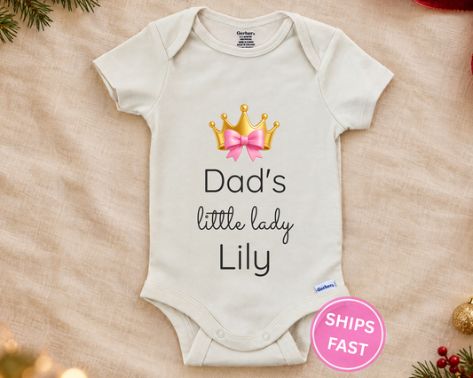 Dad's Little Lady personalized baby bodysuit with gold crown and pink bow on a cream blanket